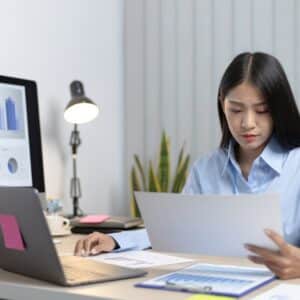woman in button down shirt looks at documents sitting at desk with laptop