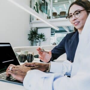 woman in blazer and glasses wits next to someone working on laptop
